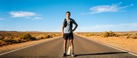 Young Caucasian male runner standing on empty desert highway wearing athletic gear, ready for training on open road with clear blue sky and arid landscape.