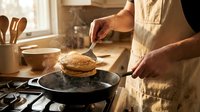 Person in apron cooking pancakes on cast iron skillet in warm sunlit kitchen. Home cooking breakfast preparation with golden morning light streaming through window.