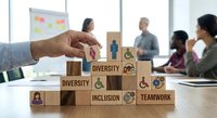Hand arranging wooden blocks with diversity and inclusion symbols in modern office meeting room with diverse business team collaborating in background.