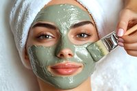 Young woman with green clay facial mask being applied with brush at spa treatment, wearing white towel on head, close up view of skincare procedure.
