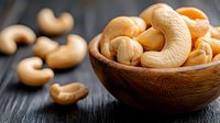 Raw cashew nuts in wooden bowl on dark rustic table, selective focus on nutritious golden nuts with scattered pieces, macro food photography.