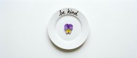 White ceramic plate with handwritten be kind message and purple pansy flower, minimalist flat lay on white background for lifestyle design.