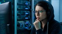 Young Asian female IT specialist with headset working in server room analyzing code on computer monitor surrounded by network equipment and data center infrastructure.