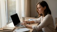 Young professional woman working on laptop at home office desk by window with coffee cup and notebook for remote work productivity.