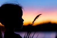 Silhouette of young girl against colorful sunset sky with lake reflection and reed grass in foreground, creating dreamy atmospheric mood at dusk.