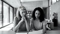 Two diverse businesswomen collaborating at modern office desk, focused on laptop screen during strategic planning meeting in contemporary workspace.