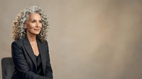 Mature woman with gray curly hair wearing black blazer, seated against neutral beige background, professional portrait for business, corporate, and executive themes.