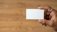 Closeup of male hand with medium skin tone holding blank white business card against wooden background for corporate branding mockup.