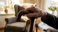 Elderly hands resting on wooden walking cane in cozy living room with armchair and natural sunlight streaming through window.