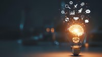 Glowing light bulb with innovation icons floating above on dark blurred background representing creative ideas, business solutions and technological advancement.