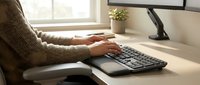 Professional woman typing on ergonomic keyboard at modern office desk with computer monitor and plant creating productive workspace atmosphere.