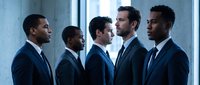 Five diverse professional businessmen in elegant dark suits standing together in modern office environment for corporate teamwork concepts.