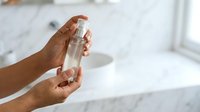 Close-up of a person with medium-dark skin tone holding a clear glass bottle of liquid, demonstrating product usage or skincare routine in bright, minimalist bathroom setting.