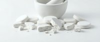 White mortar and pestle with crushed chalk, powder, and tablets scattered on white surface for pharmaceutical, cosmetic, or culinary preparation.