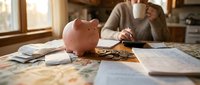 Woman planning personal finances with pink piggy bank and documents on kitchen table during morning coffee break.