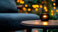 Glowing candle in dark glass holder on wooden table against blurred bokeh lights background creates cozy evening atmosphere at home.