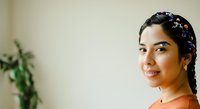 Young woman with dark hair in braids adorned with purple beads, wearing an orange top, smiling at camera in bright indoor setting with plant background.