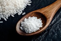 White rice grains on wooden spoon against dark stone background, macro food photography showing texture and detail of uncooked rice kernels.