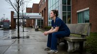 Female healthcare worker in navy scrubs sitting on concrete bench outside modern medical facility during break time reflecting on patient care responsibilities.