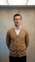Professional middle-aged Caucasian businessman in brown cardigan and white shirt standing confidently in modern office environment for corporate portraits.