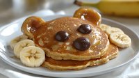 Cute bear-shaped pancake with chocolate chip eyes and nose, topped with maple syrup and served with fresh banana slices on white plate, fun breakfast food styling.