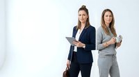 Two confident professional businesswomen standing together in modern office attire, brunette with tablet and blonde colleague in suits for corporate presentations.