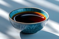 Traditional ceramic bowl with turquoise glaze pattern containing dark soy sauce, casting dramatic shadows in natural sunlight on blue surface.