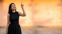 Young Hispanic woman in black dress pointing upward against golden sunset sky background for inspirational business and motivation concepts.