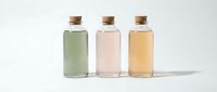 Three glass bottles with cork stoppers containing colored liquids in green, pink and yellow tones on white background for cosmetic and wellness products.