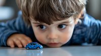 Young boy with brown hair lying down looking at small blue toy car on white surface. Child playing with miniature vehicle indoors. Childhood curiosity and imagination.