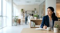 Young woman with dark hair and South Asian ethnicity, wearing professional navy blazer and white shirt, sitting at wooden desk with laptop and coffee cup in modern office environment, suitable for business, corporate, and professional themes.