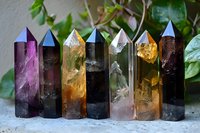 Colorful healing crystal points collection featuring amethyst, citrine, smoky quartz, and clear quartz towers arranged in row against natural green foliage background.