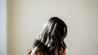 Young woman with long dark wavy hair viewed from behind in soft natural lighting against neutral wall background for wellness and lifestyle concepts.