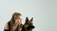 Young woman with blonde braided hair and dark makeup posing with black German Shepherd dog against neutral background for pet care, training, and animal companionship concepts.
