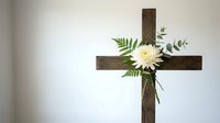 Wooden cross with white chrysanthemum flower and green fern leaves against neutral wall for memorial, funeral, Easter, or religious ceremony decoration.