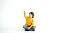 Young boy in yellow sweater sitting cross-legged pointing upward with finger on white background isolated for design use in educational materials.