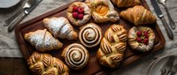 Assorted freshly baked pastries on a wooden tray, including croissants, cinnamon rolls, and Danish with fruit toppings, styled for food photography.