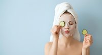 Young woman with towel on head applying facial mask holding cucumber slices for skincare routine and spa treatment at home.