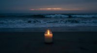 Single candle glowing on dark beach at twilight with ocean waves and dramatic sky creating peaceful meditation atmosphere for relaxation and mindfulness.