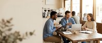 Diverse team of young professionals collaborating at wooden table during business meeting, using laptop and documents for project planning and teamwork in modern office environment.