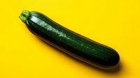 Fresh dark green zucchini squash on vibrant yellow background, dramatic lighting creates shadow effect for food photography and healthy eating concepts.