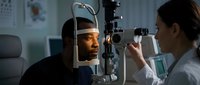 African American man receiving comprehensive eye examination from female optometrist using advanced slit lamp equipment in modern ophthalmology clinic.