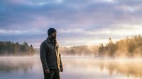 Caucasian bearded man in winter jacket and beanie stands by misty lake at golden sunrise, reflecting solitude and nature connection.