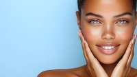Young African American woman with radiant smile and glowing skin touching face against blue background, showcasing natural beauty and skincare concept.