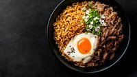 Korean beef bowl with spicy noodles, sunny side up egg, green onions and sesame seeds served in black ceramic bowl on dark background. Perfect for menu.