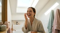 Young woman with medium skin tone applying skincare product to face in bright, modern bathroom with natural light, wearing beige robe, suitable for beauty, wellness and self-care content.