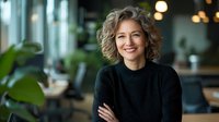 Confident mature businesswoman with curly blonde hair wearing black sweater, arms crossed, standing in modern office space with natural light and plants.