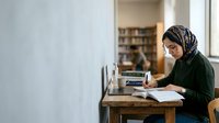 Woman wearing hijab studying at wooden desk with laptop and books in library setting.