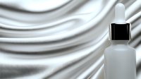 Frosted glass dropper bottle with black cap on luxurious white silk fabric background. Mockup for cosmetic skincare product presentation.