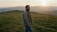 Man with beard wearing casual jacket standing in rolling green hills at sunset, looking toward distant mountains and golden sky light.